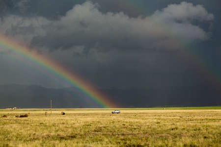 Sun illuminating prairie grassland with dark and forbidding thunderclouds and downpour with van riding along lonely country road and rainbow ending over the vehicleの写真素材
