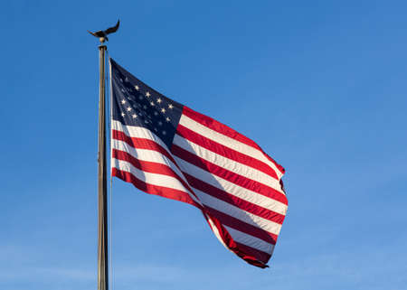Wind blows the USA flag of stars and stripes with eagle flagpole against blue sky late in the...