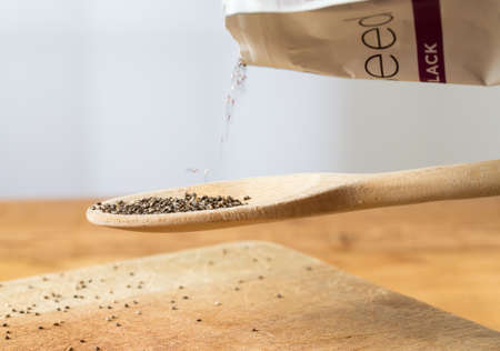 Superfood known as black chia seeds are poured from packet into a wooden backing spoon over wood bread board on kitchen tableの写真素材