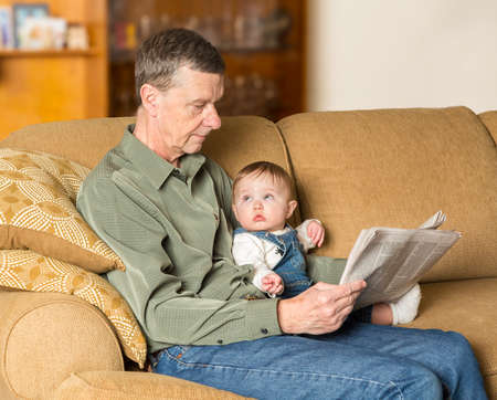 Young caucasian baby girl looking up at grandfather reading newspaper on settee in family roomの写真素材