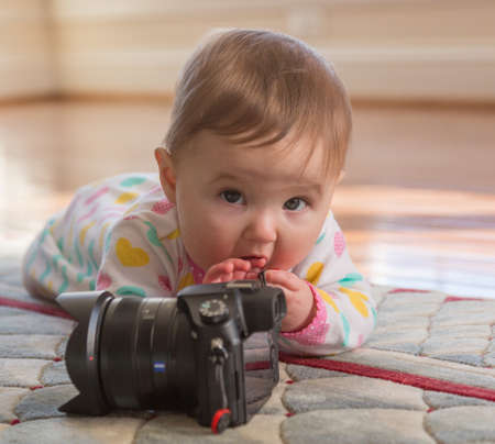 Young caucasian baby girl lying on carpet or rug and playing with an expensive camera as a budding...