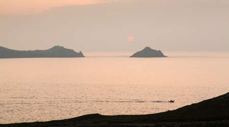 Small boat races back to port by headland overlooking the coast at sunset at Port Quin, Cornwall, England, United Kingdomの写真素材