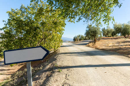 Blank road sign by side of dry dusty road with olive trees and mountains in the distance