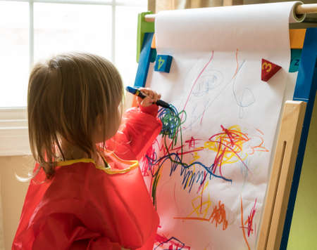 Young two year old girl drawing with crayon on paper mounted on an easel during playtimeの写真素材