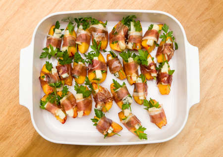 White plate full of home made appetizers with peach slice and gorgonzola cheese wrapped by strip of prosciutto hamの写真素材