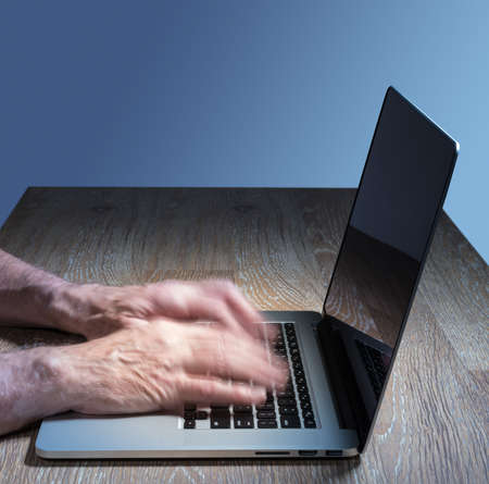 Senior caucasian adult man types on modern keyboard of laptop or ultrabook on wooden desk with copy...