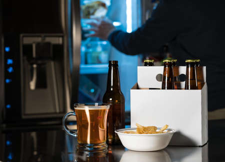 Six pack of brown beer bottles in plain white cardboard carrier with mug of ale on stainless steel kitchen or bar counter. Open fridge or refrigerator out of focus in rear.の写真素材