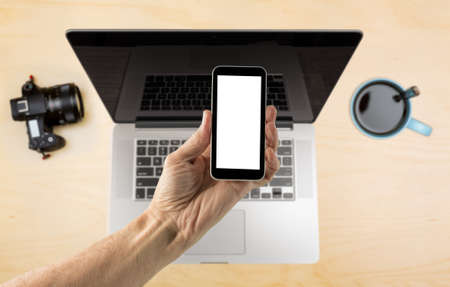 Male hand holding a smartphone with a blank screen over organized desk with laptop blurred in...