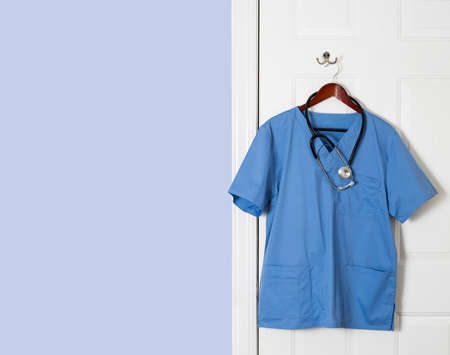 Blue medical scrubs uniform shirt hanging on a hook on back of door with stethoscope with copy space for messageの写真素材