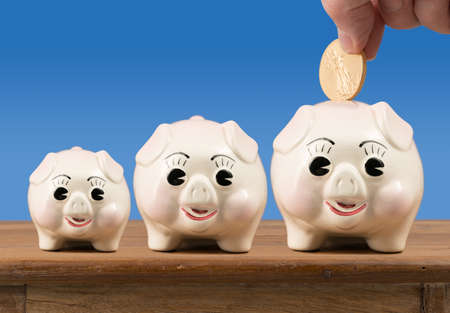 Three piggybanks in small, medium and large sizes sitting on a wooden shelf or table with gold coin being inserted into largest oneの写真素材
