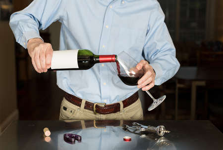 Senior caucasian man standing in kitchen pouring from bottle of red wine with a blank label for copy spaceの写真素材