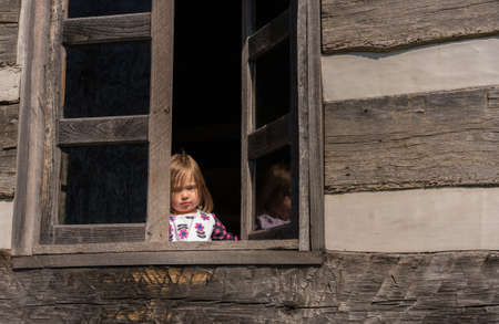 Small and lonely caucasian baby girl or toddler looking out of old wooden cabin window to suggest...