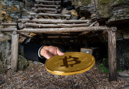 Miner engaged in mining for new bitcoins holds out a coin out of the entrance to an old coal mine