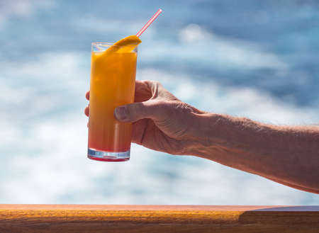 Senior caucasian adult man with tequila sunrise in glass on teak balcony rail of ocean cruise ship