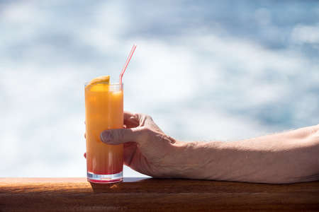 Senior caucasian adult man with tequila sunrise in glass on teak balcony rail of ocean cruise shipの写真素材