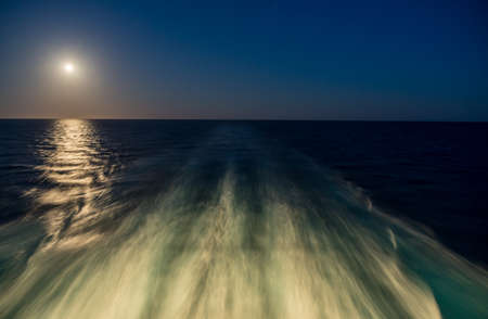 Moon rising over wake and waves of cruise ship at sea with concept of leaving or starting anewの写真素材