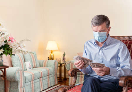 Senior caucasian man counting some US dollar bills. He is wearing face mask to prevent coronavirus. Composite into home backgroundの写真素材