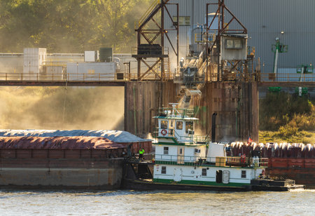Tugboat pushing freight barges past grain loading dock with dust illuminated by the sun in East St...