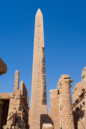 Ancient Egyptian obelisk covered in hieroglyphs stands tall against a clear blue sky, surrounded by ruins of Karnak templeの写真素材
