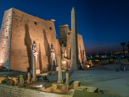 Massive stone wall and standing and seated statues with obelisk in Luxor Temple in Egypt illuminated at nightの写真素材