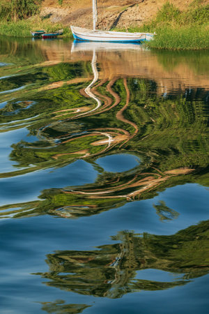 A small sailboat floats on a calm river Nile, its reflection distorted by gentle ripples creating an abstract pattern on the waters surface.の写真素材