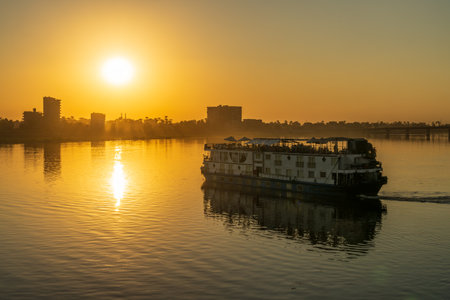 A cruise ship sails along the Nile River at sunset, reflecting the golden light. Buildings are silhouettedの写真素材