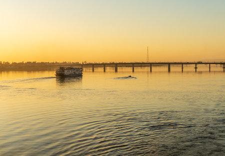 A older cruise ship sails along the Nile River at sunset, reflecting the golden light. Buildings are silhouettedの写真素材