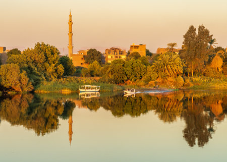 Tranquil riverside town of Naqadah in Egypt, with traditional buildings and lush vegetation, with boat on riverの写真素材