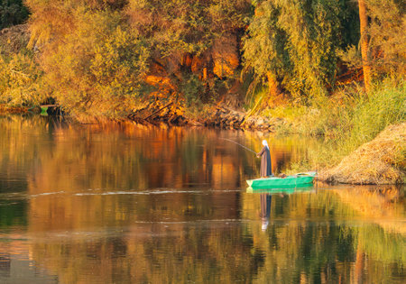 Man in traditional galabia fishing from small green rowboat by banks of river Nileの写真素材