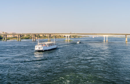 River cruise boat sailing upstream from modern bridge connecting east and west banks of the Nile at...