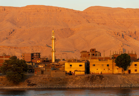 Minaret at sunset with home construction and large sand mountain of Sahara desert by banks of Nile Riverの写真素材
