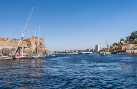 Skyline of Aswan in distance with Felucca boat docked by Ruins of Abu and many river cruise boats...