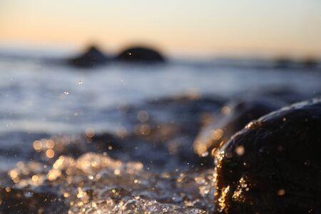 Closeup photo of sea waves braking on a wet seashore stones summer sunset, water splashes , sun rays, reflections on water drops.の写真素材