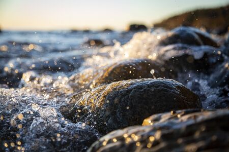 Closeup photo of sea waves braking on a wet seashore stones summer sunset, water splashes , sun rays, reflections on water drops.の写真素材