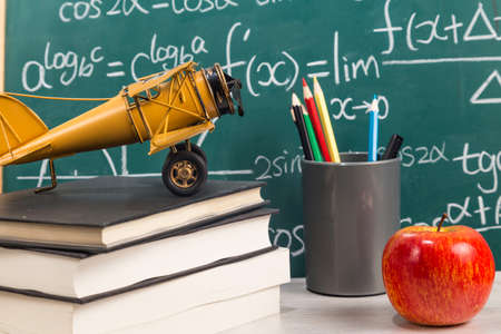 Apple, books, aircraft toy and pencils on desk with blackboard in the background