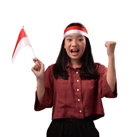 Excited young Asian woman cheering with an Indonesian flag and wearing a red and white headband, celebrating national pride, sports victory, or Independence Day. Isolated on white.の写真素材