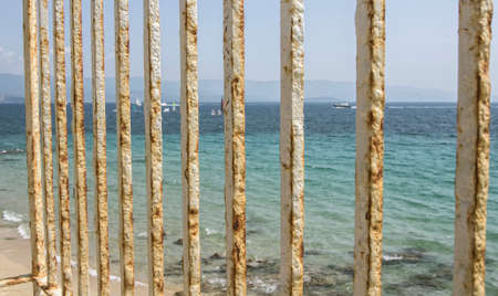 An old rusty white painted fence with the blue sky and beach with boats in the background.の写真素材