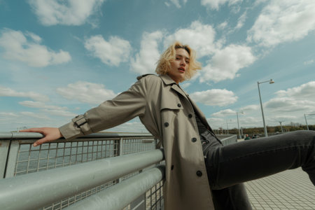 A stylish young woman with blonde hair and a grey trench coat leans on a railing. The background features a dramatic cloudy sky suggesting an urban outdoor setting.の素材