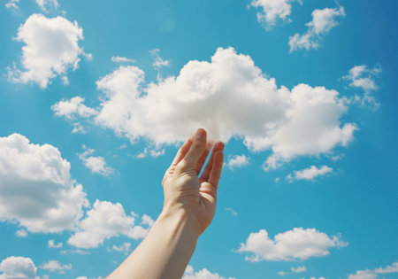 A hand reaches up to touch a soft, white cloud against a vibrant, clear blue sky filled with scattered clouds.の素材