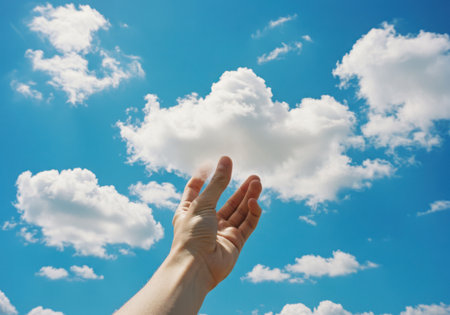 A person's hand reaches up towards fluffy white clouds against a bright blue sky, symbolizing aspiration and dreams.の素材