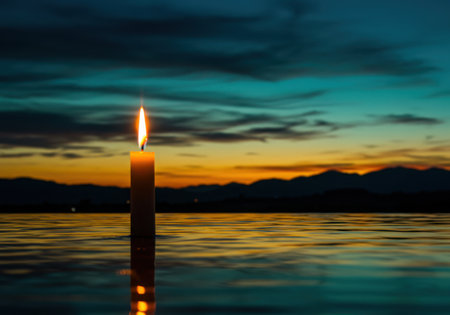 A single lit candle stands tall in the foreground, its flame reflected on the calm water as a vibrant sunset paints the sky.の素材