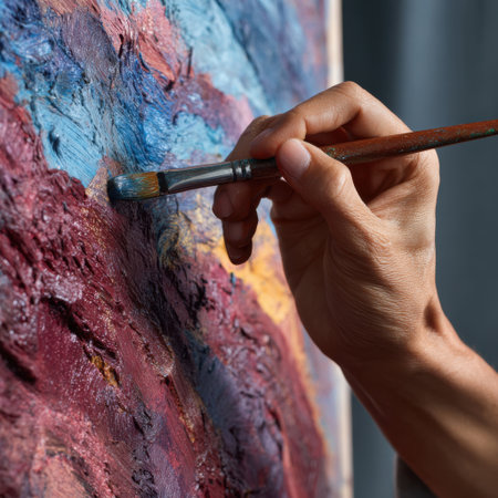 Close-up of an artist's hand holding a brush, adding vibrant, textured strokes to a colorful abstract painting on canvas.の素材
