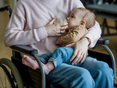 A mother in a wheelchair lovingly feeds her baby from a bottle, highlighting care and family connection.の素材