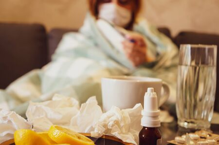 The concept of the common cold or seasonal flu. A young girl in a mask looks at a thermometer sitting on a sofa wrapped in a blanket near a table with medicines. Drinks a hot healing drink. Quarantineの写真素材