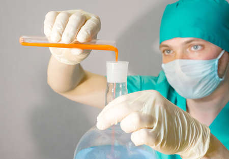 A doctor in a medical mask and latex gloves looks at the tests in a flask. holding a flask with red...