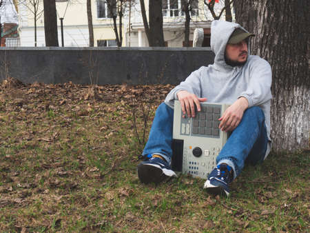 A hip-hop beatmaker sits on the grass in a park under a tree holding an old-fashioned '90s drum...
