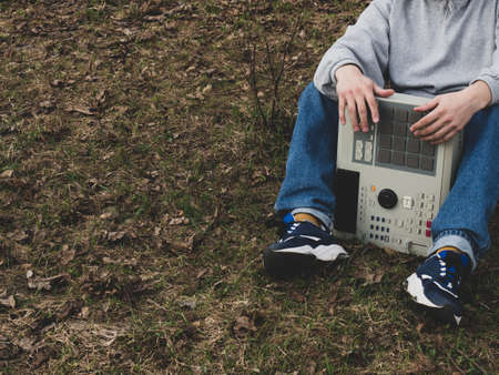 A hip-hop beatmaker sits on the grass in a park under a tree holding an old-fashioned '90s drum machine. Retro Digital Musical Instrument for Hip Hop Producers and Beatmakersの写真素材