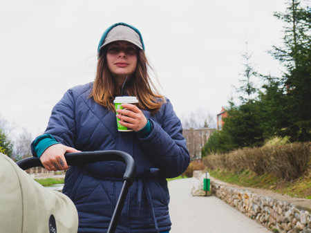 A woman in a baseball cap walks with a stroller in the park and drinks coffee