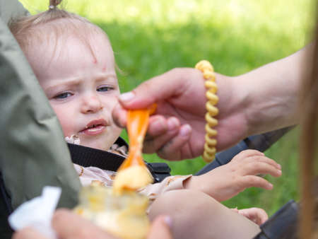a young mother feeds a baby on the street in a stroller with a spoon from a jar of mashed potatoes...