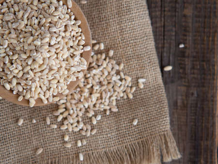 raw pearl barley in a wooden plate and a wooden spoon next to it, spilling on a dark wooden tableの写真素材